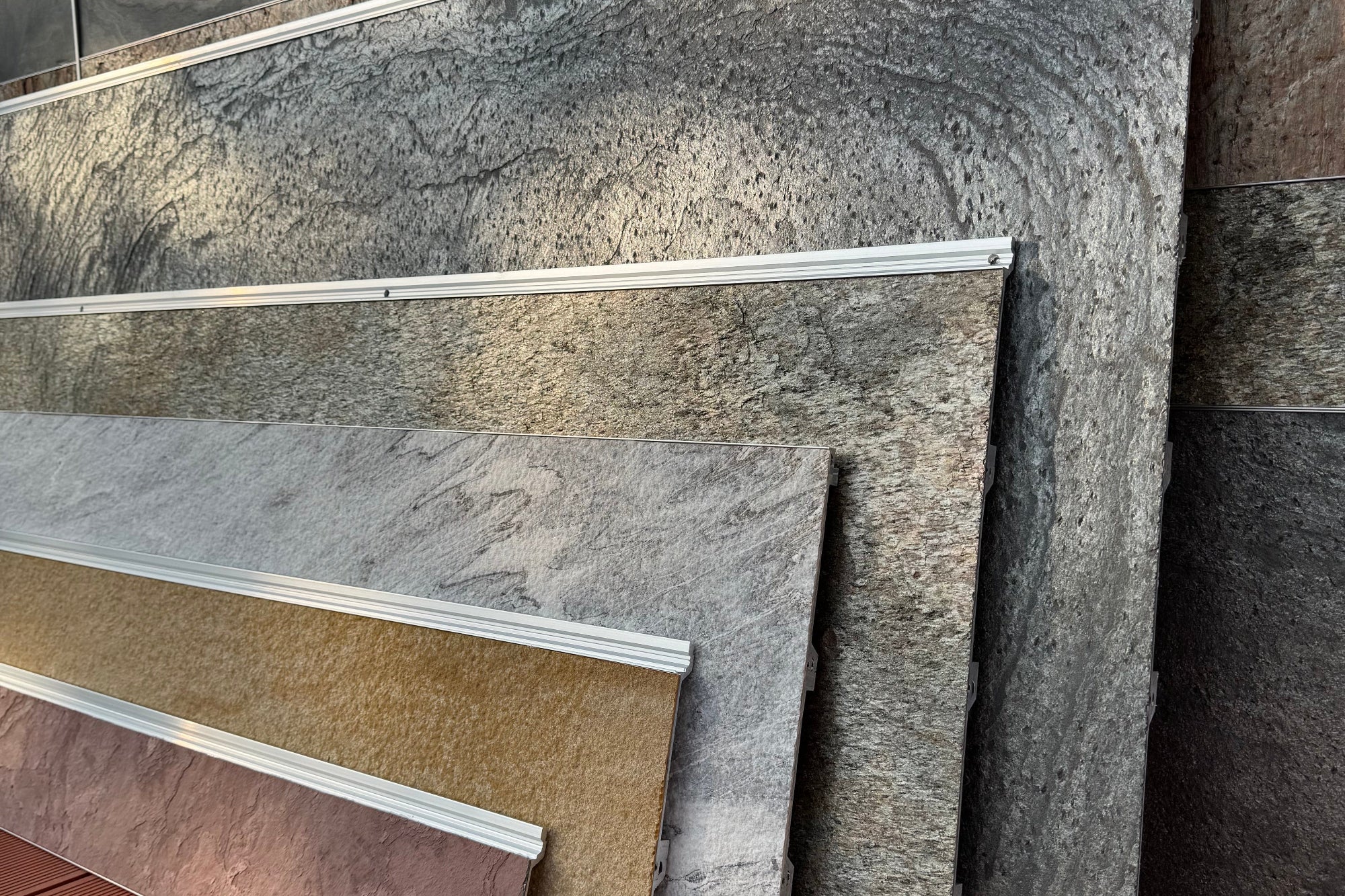 A stack of large stone-look exterior wall panels in gray, brown, and beige tones, leaning against a matching stone wall on an orange-brown deck.