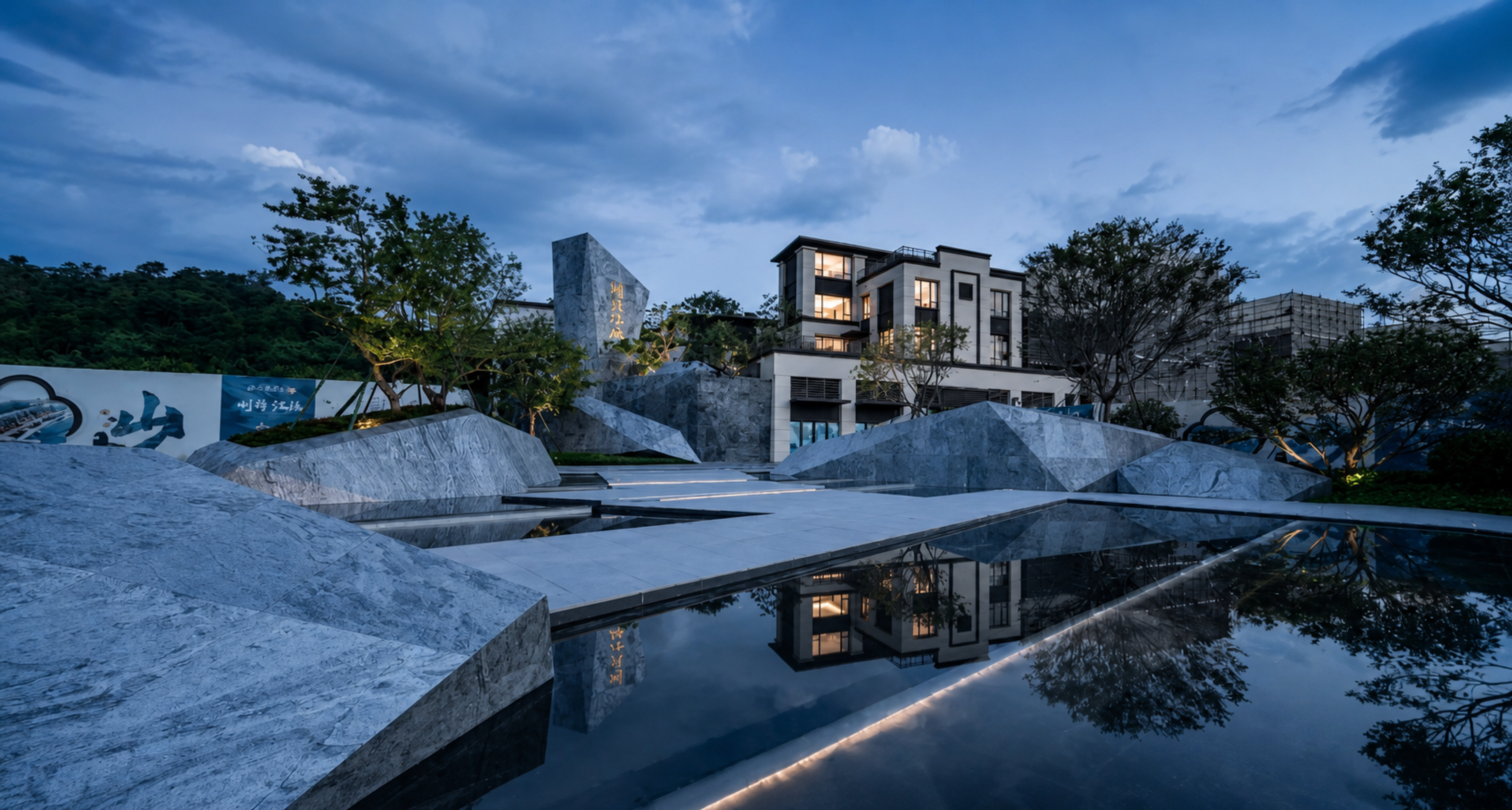 Modern multi-story building with lit windows set among trees and stone terraces, reflected in a dark water feature at dusk under a blue evening sky.