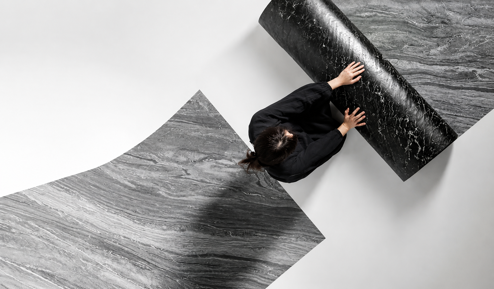 A person in a black outfit is shown from above, holding and unrolling a sheet of dark gray marble-look wallpaper or adhesive flooring against a clean white background.