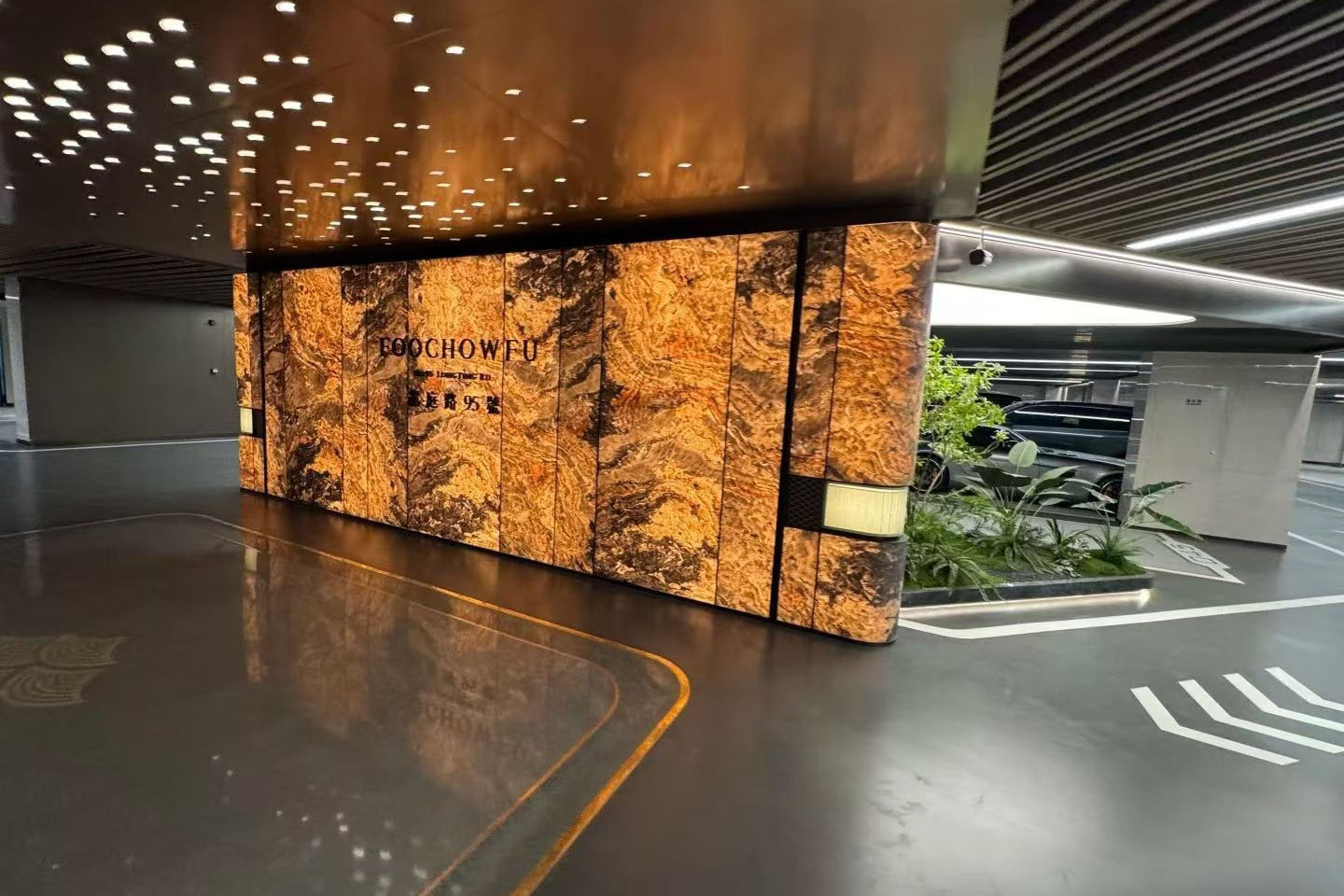 A modern interior space with a large illuminated stone wall panel displaying the brand name 'BAOCHOWEI' in gold lettering. The wall features textured golden-brown and black marbled stone panels, set against a dark gray floor and ceiling with decorative lighting.