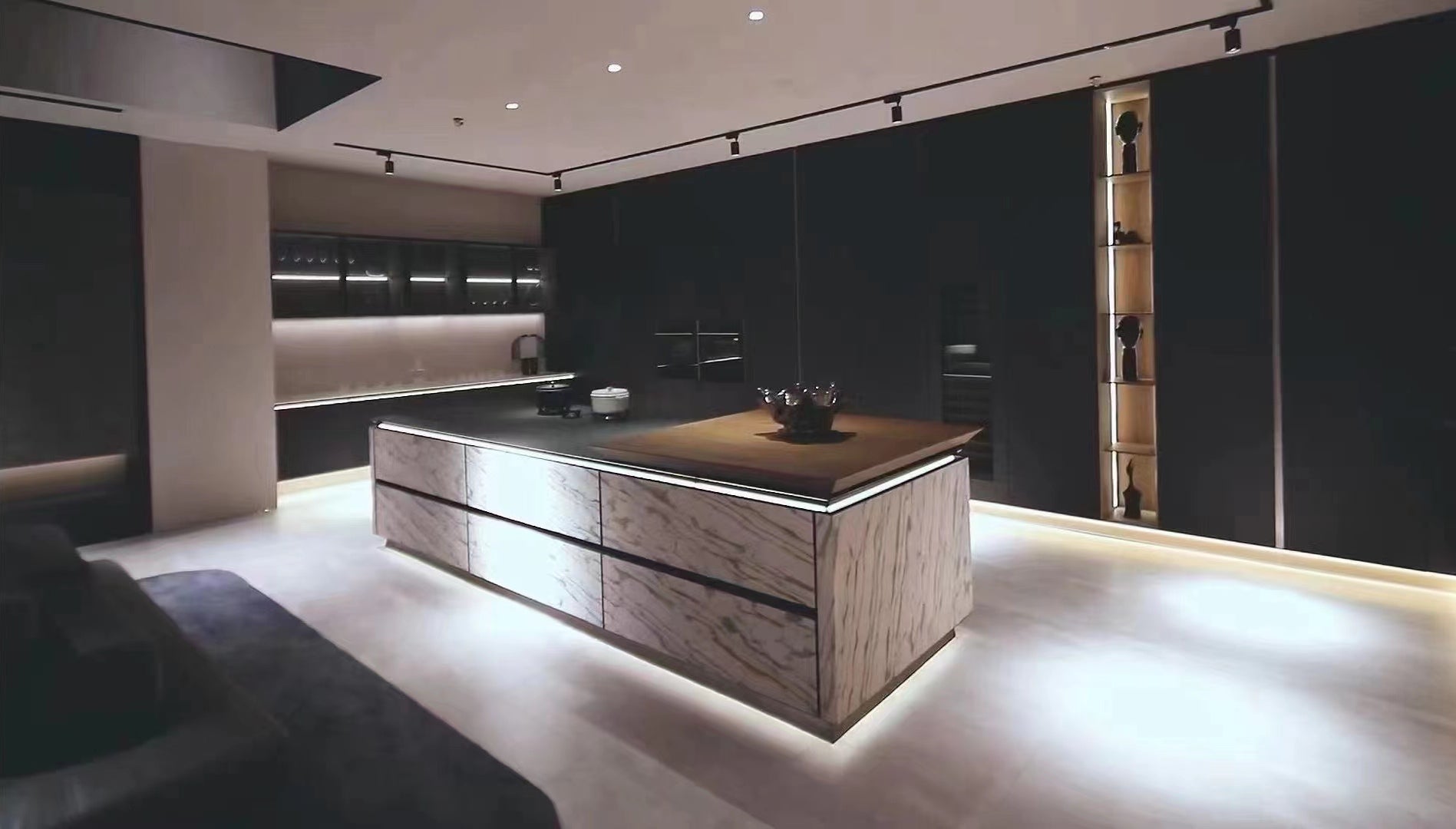 A modern kitchen island with a light wood grain front and a dark countertop, set in a minimalist kitchen with black cabinetry and warm under-cabinet lighting.